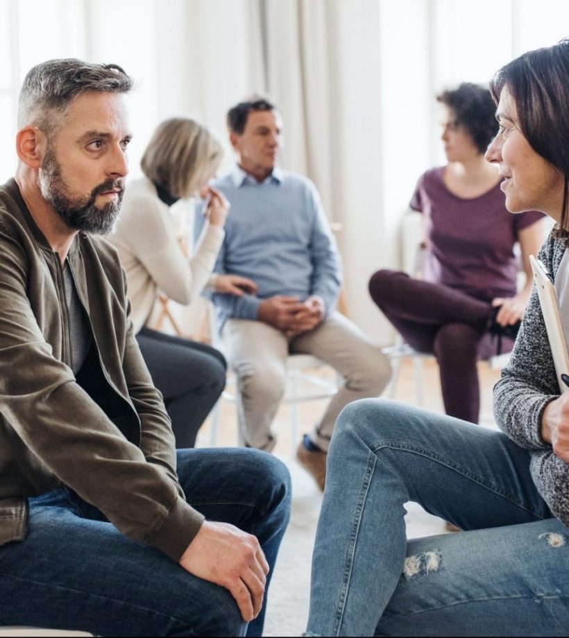 senior-counselor-with-clipboard-talking-to-a-man-during-group-therapy--e1625973530854-pf2n5igpmfljfnk0l1fnk2u5lvp83rcletgm4z0k4w
